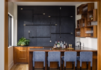 02222022-noble-112-Edit Sophisticated home bar in a custom-built Kansas residence, featuring an elegant black textured backsplash with unique pipe lighting, rich wooden cabinetry with neatly organized glassware, a polished countertop with a selection of fine spirits, and comfortable blue fabric bar stools, creating a luxurious space for entertaining.