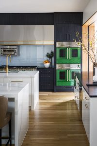 Custom kitchen with green wall ovens, black and white cabinetry and countertops, gold hardware and wood floors