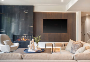 A chic and contemporary living room design featuring a cozy fireplace, sleek dark wood paneling, a large flat-screen TV, comfortable beige sectional sofa with plush pillows, a unique modern armchair, and a stylish round coffee table with books and decorative items, located in a custom-built home in Loch Lloyd, Missouri.