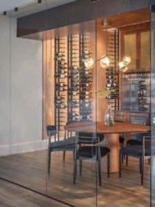 Wine cellar and bar in custom home in MO Sophisticated dining area with wine storage in a Loch Lloyd, Missouri custom-built home, featuring a floor-to-ceiling wine rack displaying a diverse collection, an oval wooden dining table flanked by sleek black chairs, and artistic pendant lights casting a warm ambiance, all enclosed by elegant glass walls enhancing the room's open and airy feel.