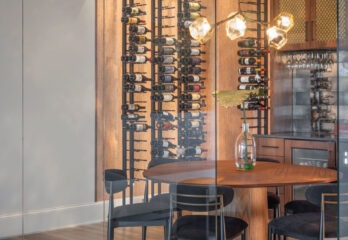 Sophisticated dining area with wine storage in a Loch Lloyd, Missouri custom-built home, featuring a floor-to-ceiling wine rack displaying a diverse collection, an oval wooden dining table flanked by sleek black chairs, and artistic pendant lights casting a warm ambiance, all enclosed by elegant glass walls enhancing the room's open and airy feel.