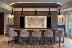 Elegant home bar in Loch Lloyd Missouri Elegant home bar in a custom-built Missouri residence featuring a curved dark wood bar with plaid upholstered stools, a striking backlit onyx feature wall, chic black pendant lights, and sophisticated accents including a round side table and a cozy blue corner seat, all set against a backdrop of rich wooden tones and soft ambient lighting.