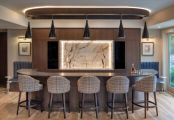 Elegant home bar in a custom-built Missouri residence featuring a curved dark wood bar with plaid upholstered stools, a striking backlit onyx feature wall, chic black pendant lights, and sophisticated accents including a round side table and a cozy blue corner seat, all set against a backdrop of rich wooden tones and soft ambient lighting.